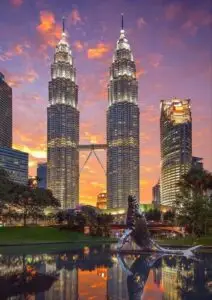 Petronas Twin Towers illuminated at sunset in Kuala Lumpur, Malaysia, reflected in a water feature surrounded by greenery.