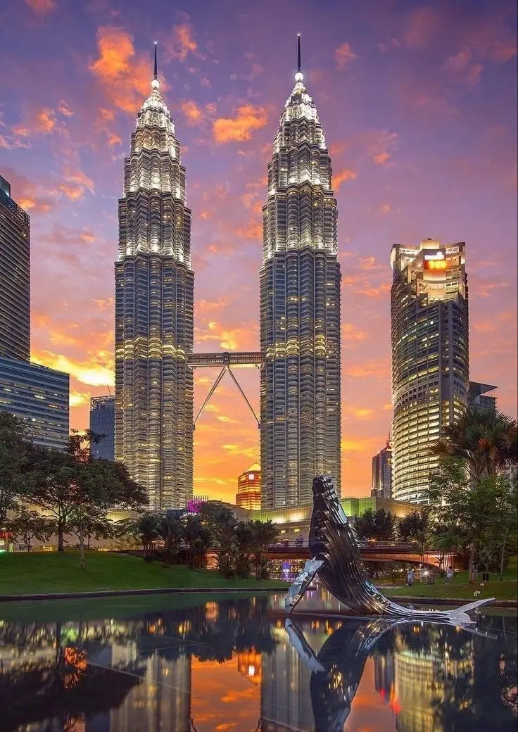 Petronas Twin Towers illuminated at sunset in Kuala Lumpur, Malaysia, reflected in a water feature surrounded by greenery.