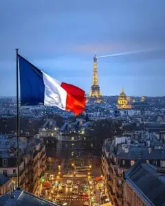 French flag waving over Paris with the illuminated Eiffel Tower and city skyline at dusk.