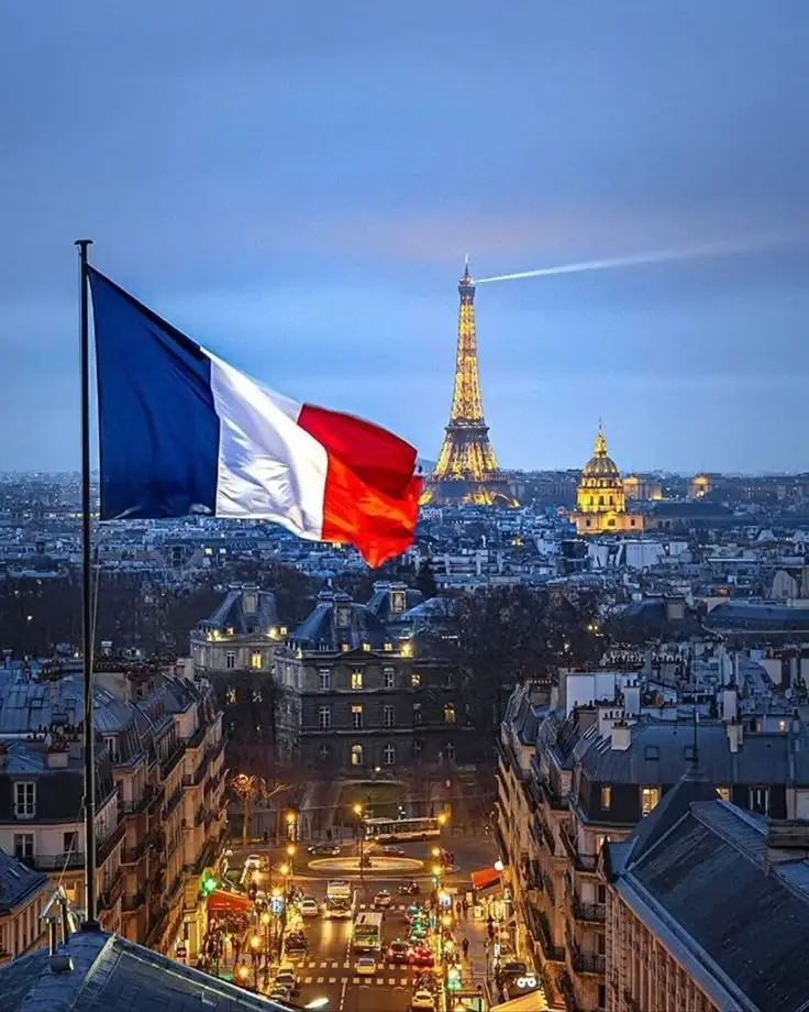 French flag waving over Paris with the illuminated Eiffel Tower and city skyline at dusk.