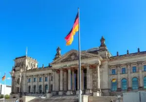 German flag flying in front of the Reichstag building in Berlin under a clear blue sky.