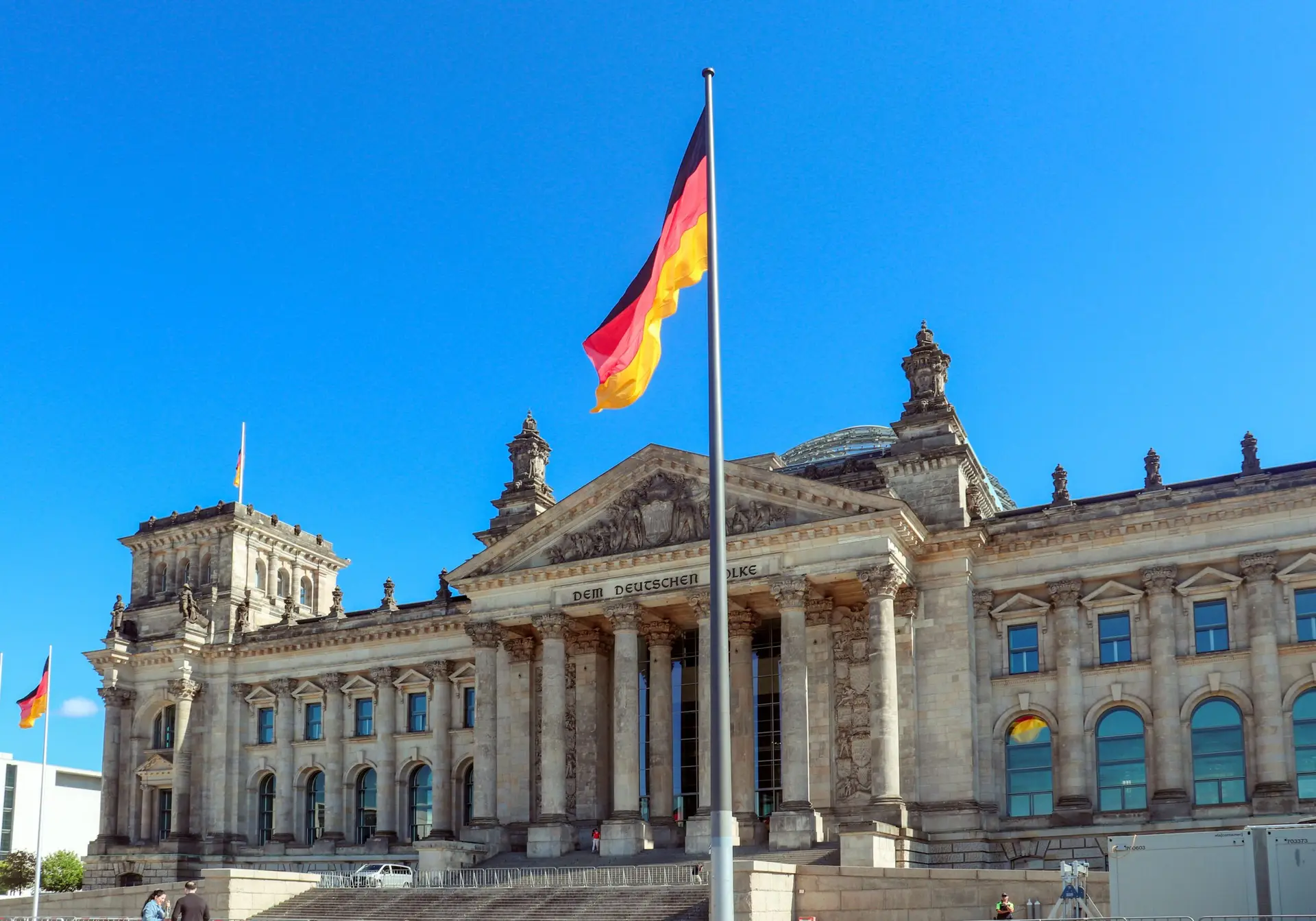 German flag flying in front of the Reichstag building in Berlin under a clear blue sky.