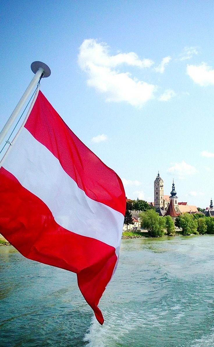 Austrian flag waving over a river with a historic riverside town and church in the background.