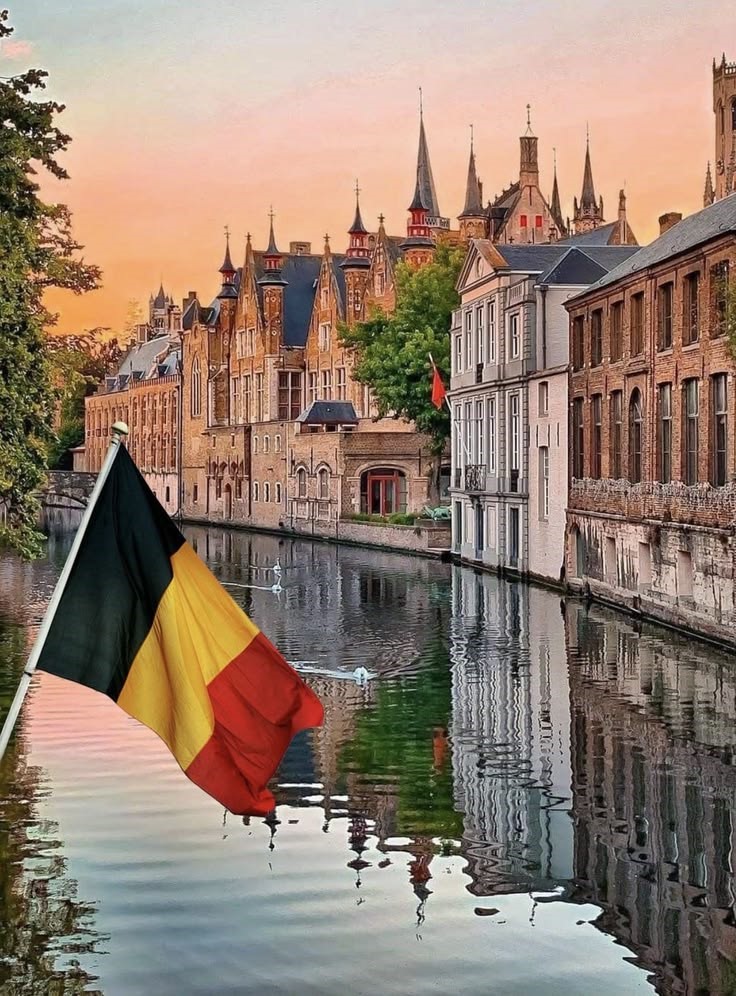 Scenic view of a historic canal in Bruges, Belgium, with traditional medieval buildings, church spires, and a Belgian flag reflected in the water.